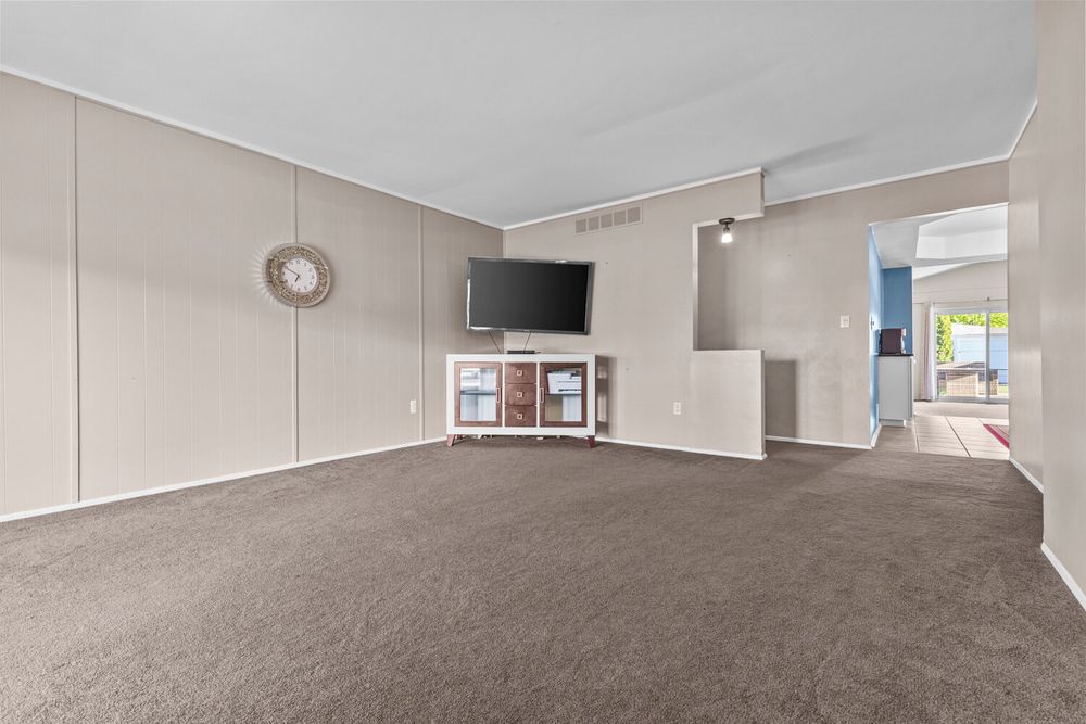 Spacious living room with gray carpet, wall paneling, mounted TV on white media console, and open view to adjacent room.