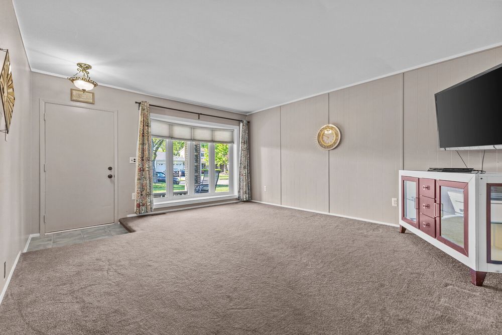 Bright living room entry with large front door, gray carpet, crown molding, wall sconce, and white TV console.