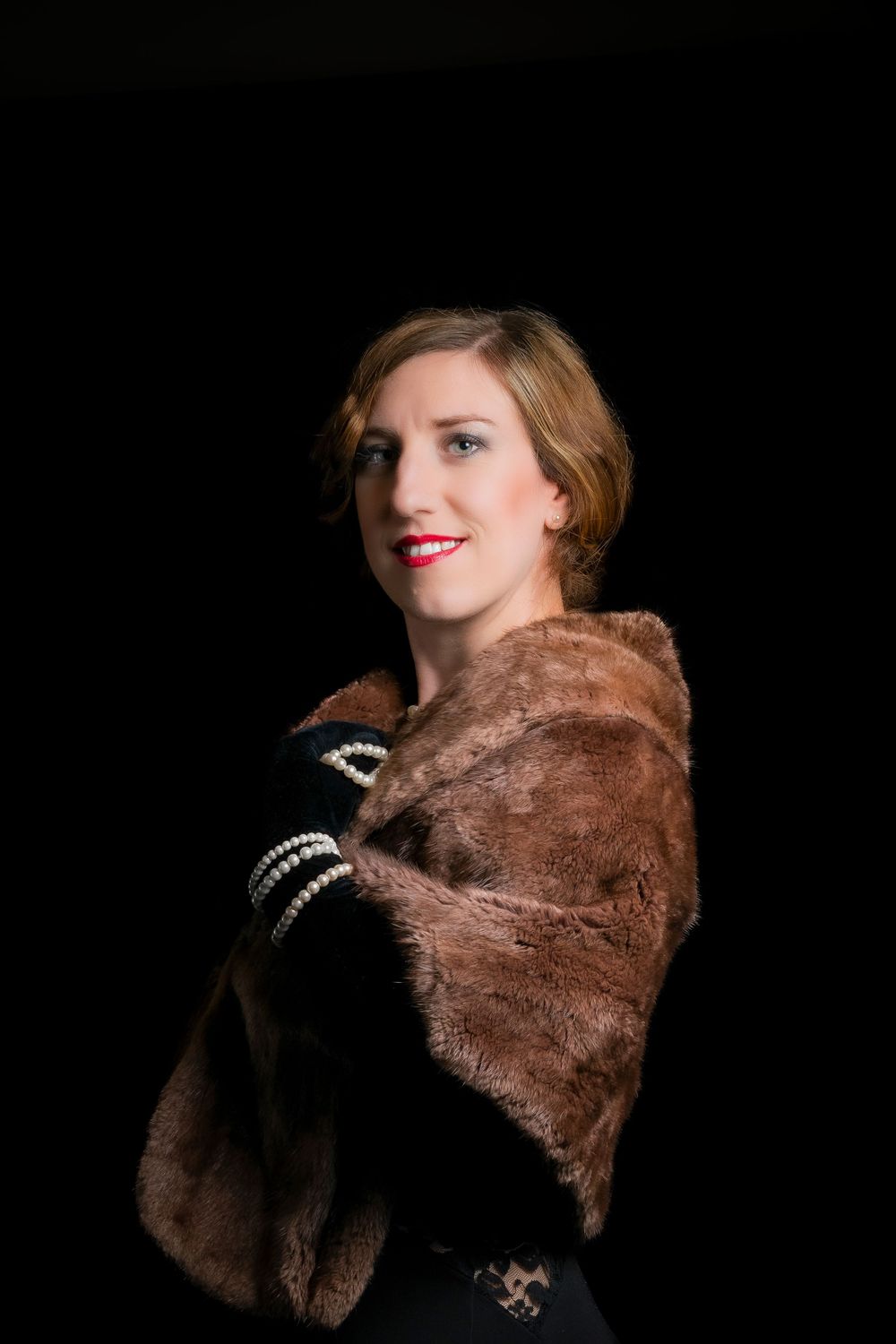 Elegant woman wearing brown fur stole and pearl bracelet posing against dark background.
