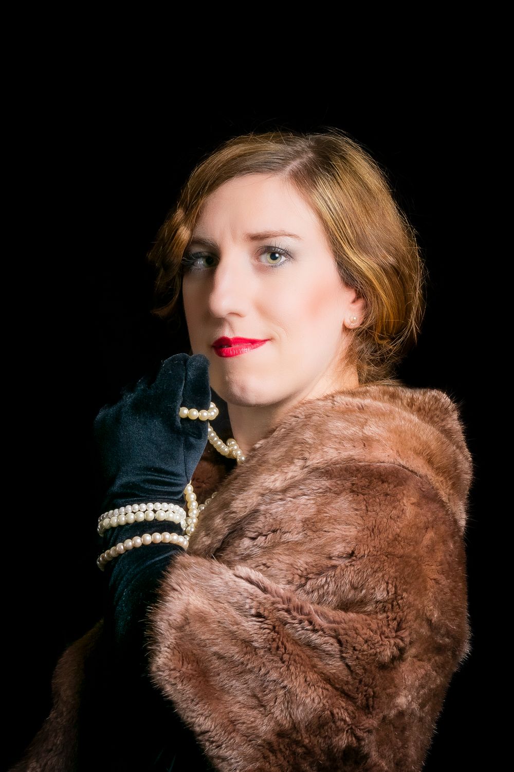 Vintage-styled woman in fur coat and black gloves holding pearl necklace with red lips on dark background.