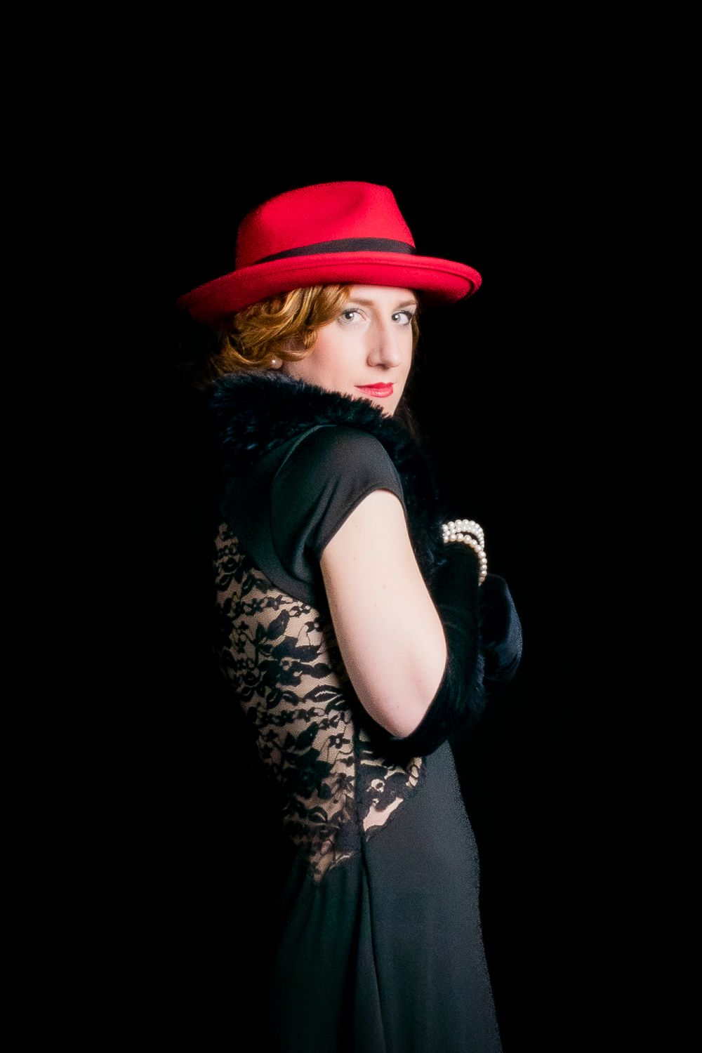 Woman in red fedora hat and black lace dress with gloves poses elegantly against dark background.