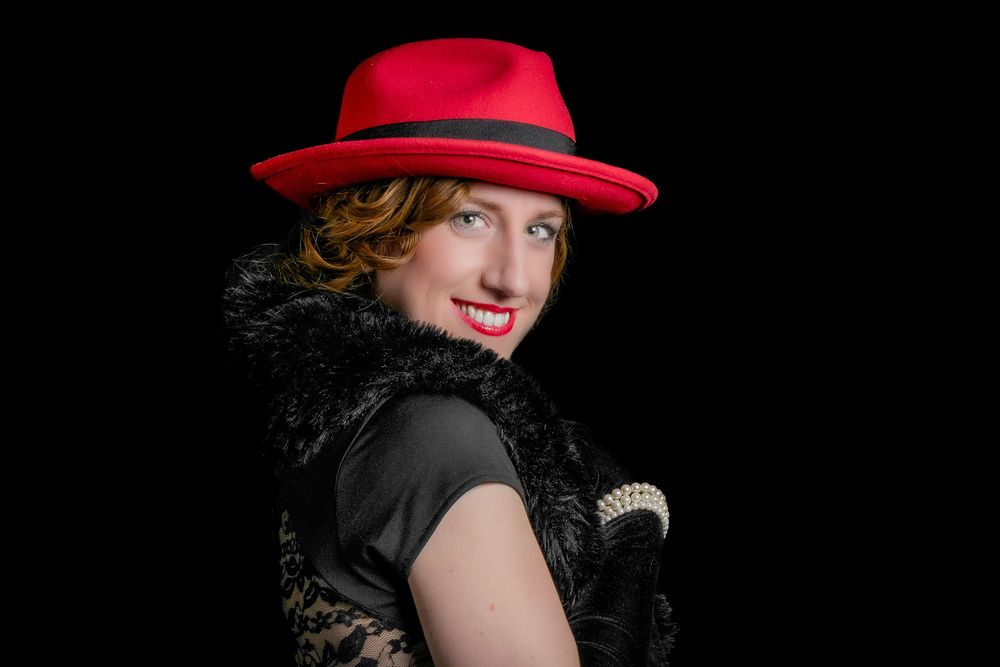 Woman in red fedora hat and black lace dress with feather boa smiles over shoulder.