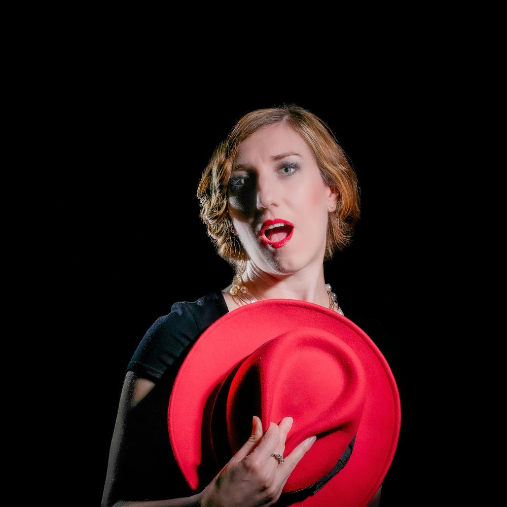 Elegant woman in black dress holding a red fedora hat with red lips and vintage-style wavy hair on dark background.