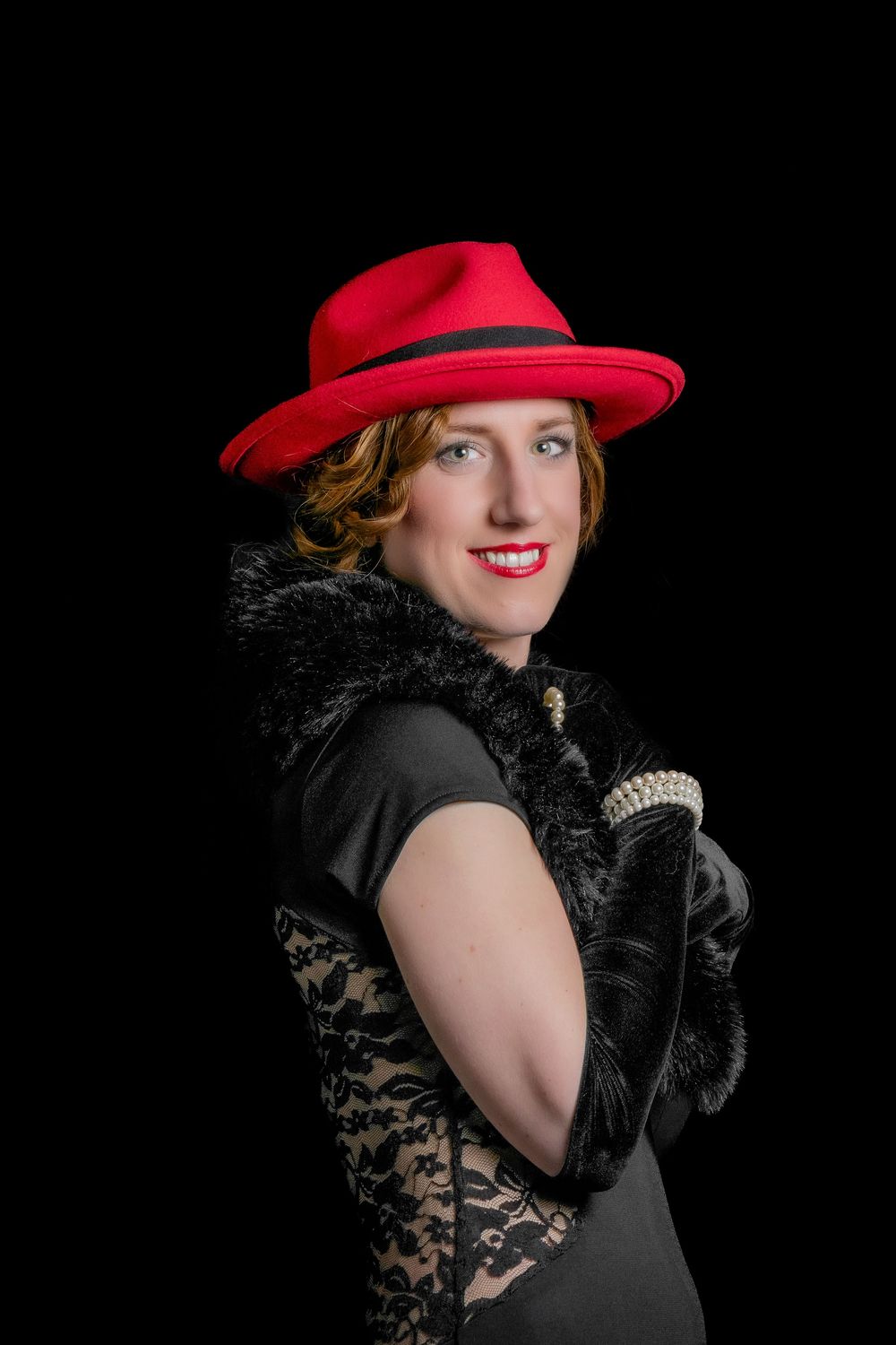Woman in red fedora hat, black lace dress, fur boa, and pearl gloves poses elegantly against dark background.
