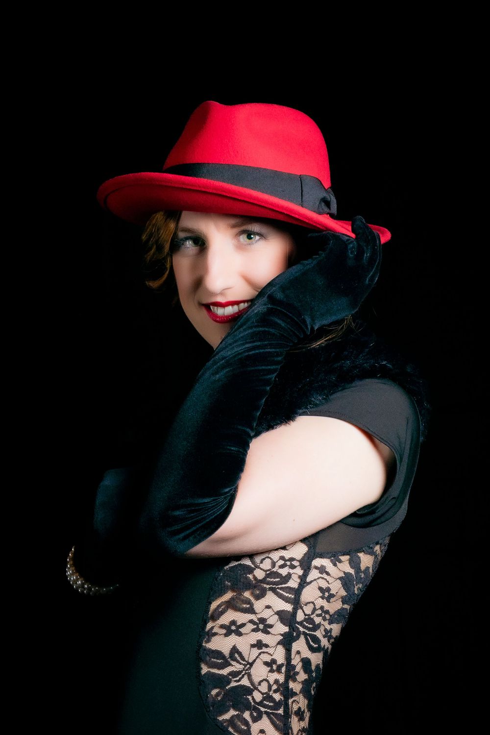 Woman in red fedora hat and black lace dress with velvet gloves posing elegantly against dark background.
