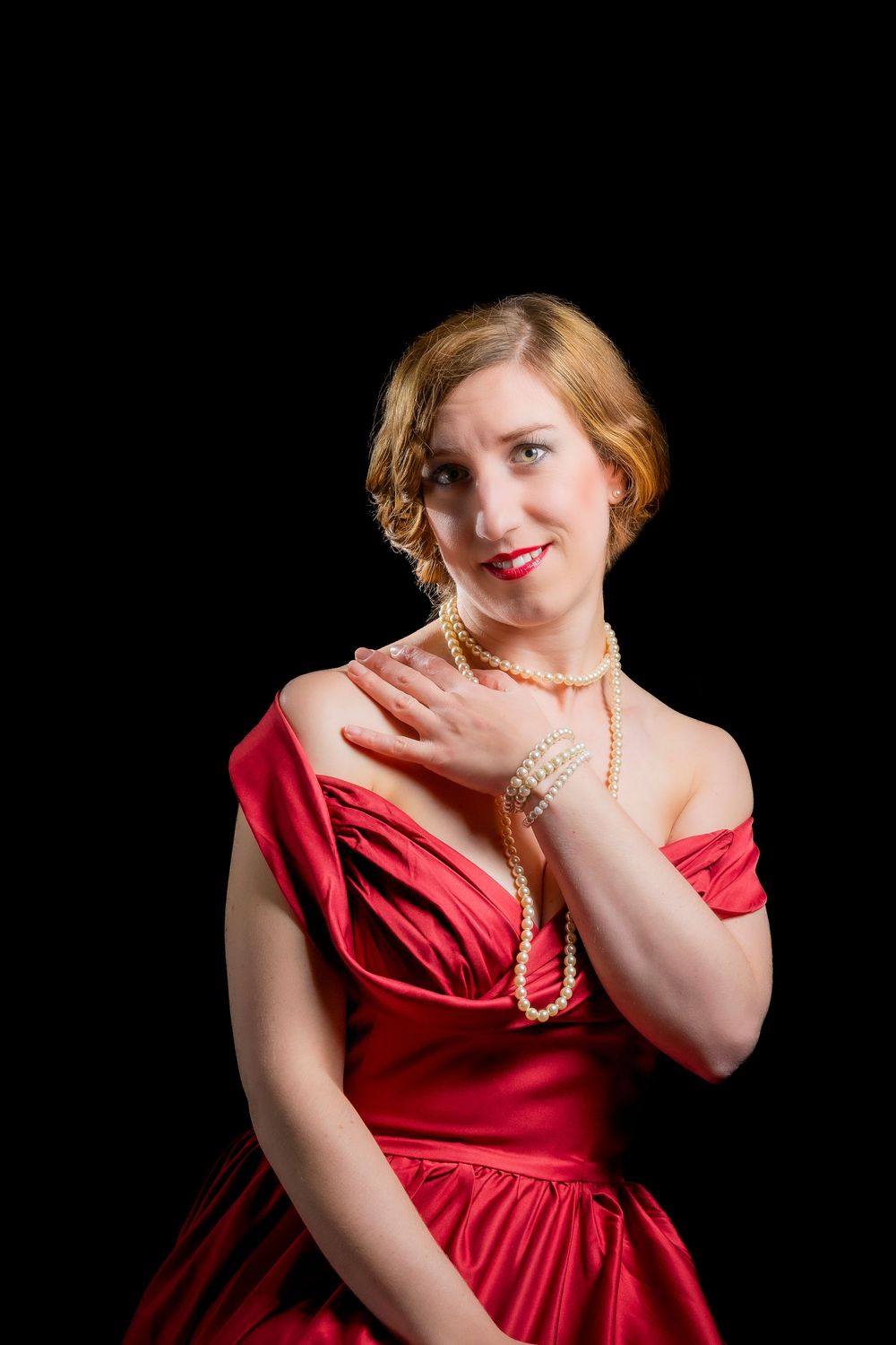 Woman in red satin off-shoulder gown with pearl necklace and bracelet posing against black background.