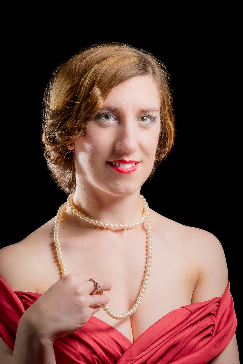 Elegant woman in red off-shoulder gown wearing pearl necklace and ring, smiling against black background.