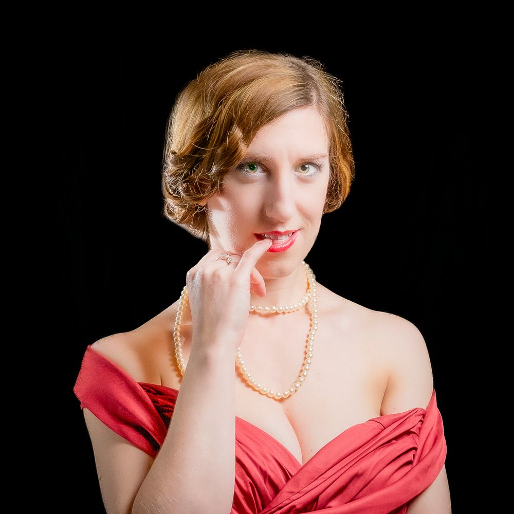 Woman in red off-shoulder dress with pearl necklace posing thoughtfully against black background.