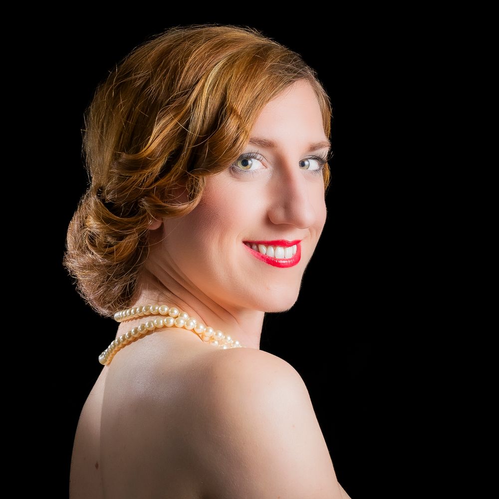Elegant woman with pearl necklace and updo hairstyle, wearing red lipstick, posed against black background.