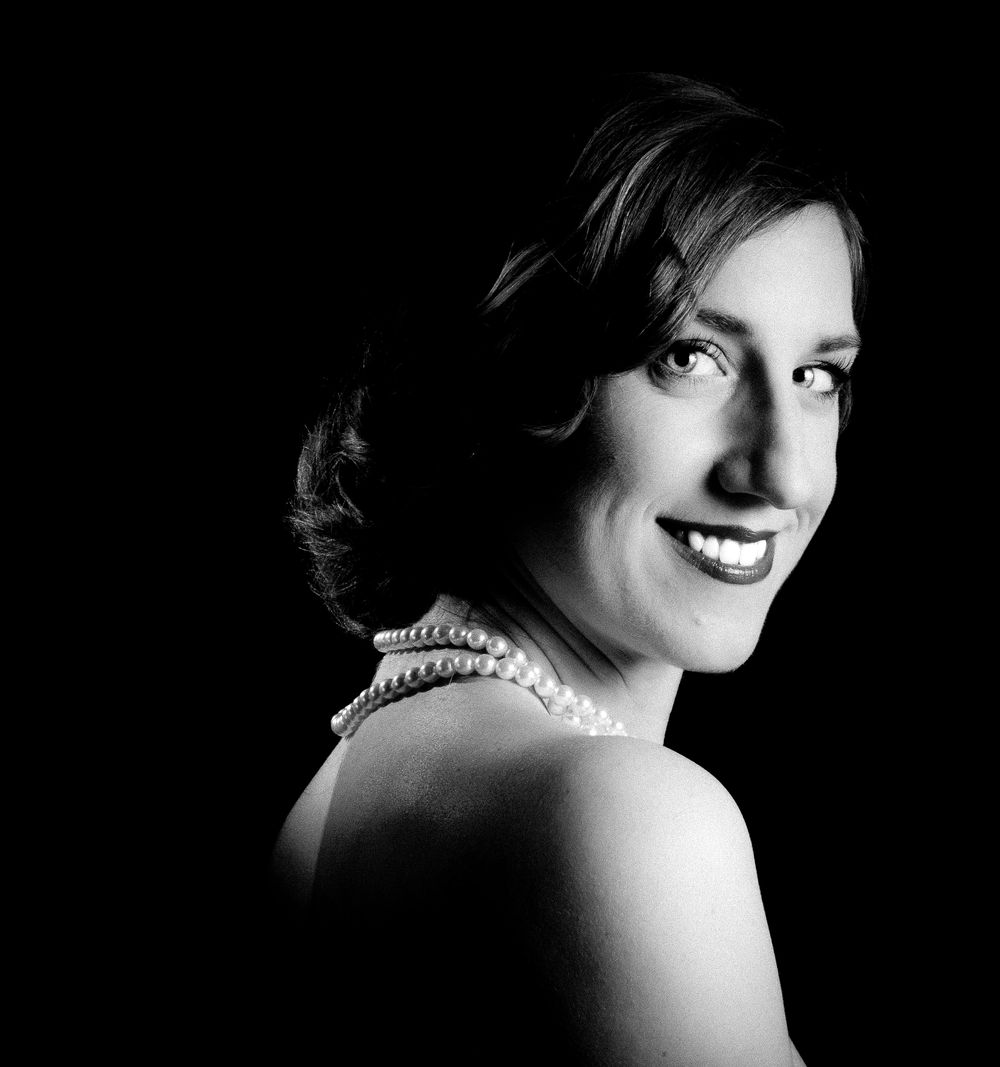 Black and white portrait of a smiling woman wearing a pearl necklace, elegant bare shoulders against dark background.
