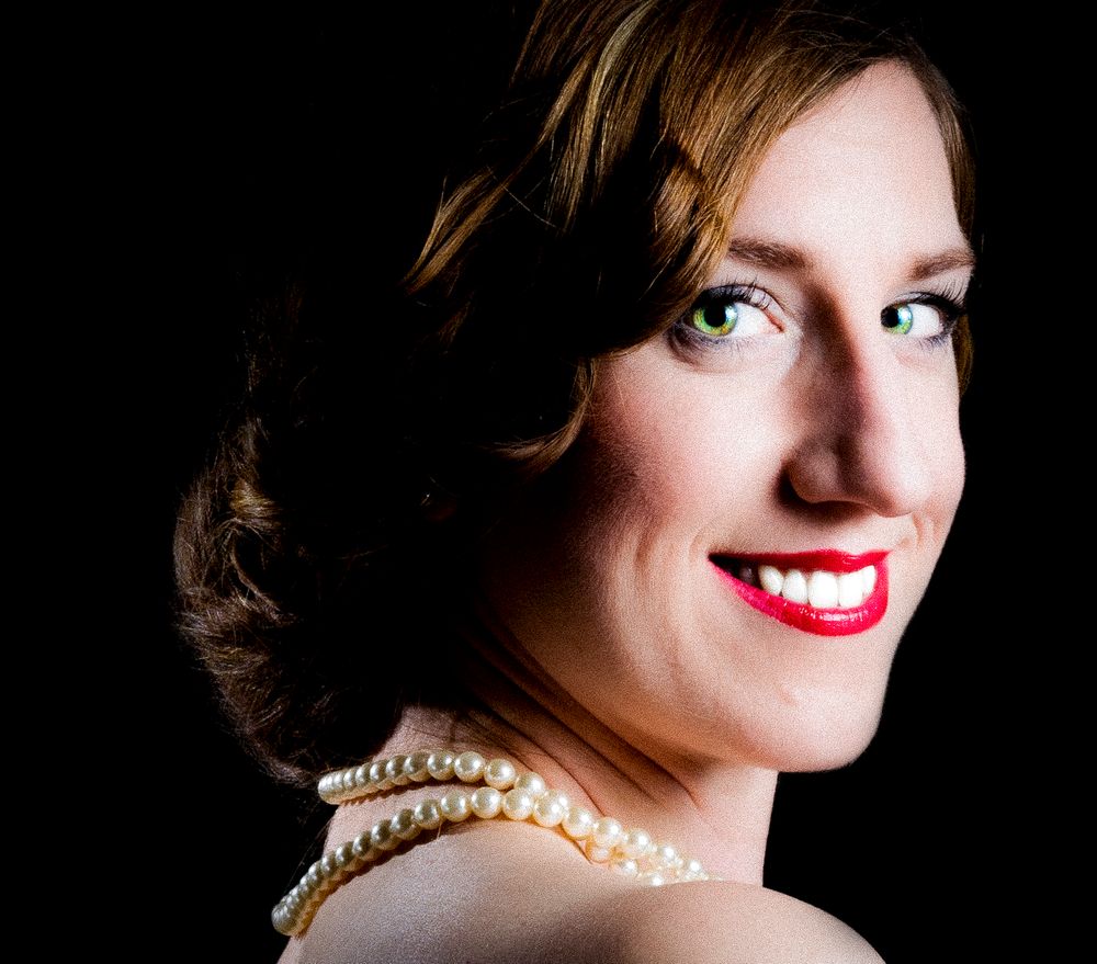 Elegant woman with green eyes, pearl necklace, red lips, and updo hairstyle posing against dark background.