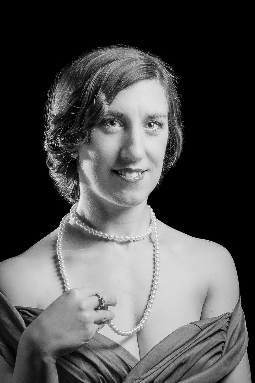 Black and white portrait of a young woman wearing pearl necklace and elegant gown, smiling against dark background.