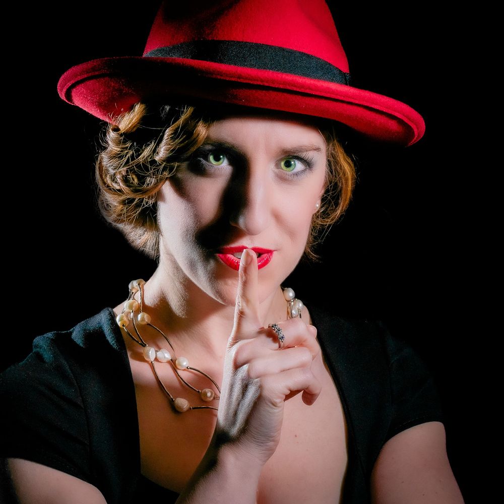 Woman in black top with red fedora hat making silence gesture wearing pearl necklace.