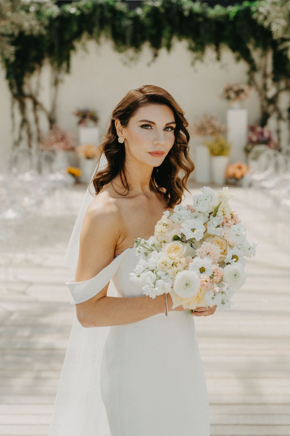 Elegant bride in off-shoulder white gown holding a lush bouquet of white and blush flowers at an outdoor wedding venue.