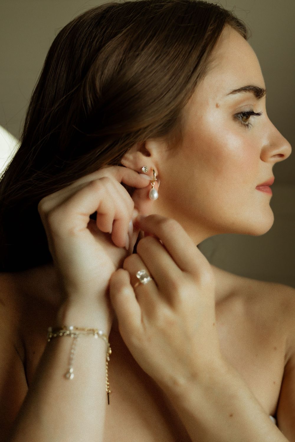 Woman putting on pearl drop earrings with gold bracelet and ring, elegant jewelry styling.