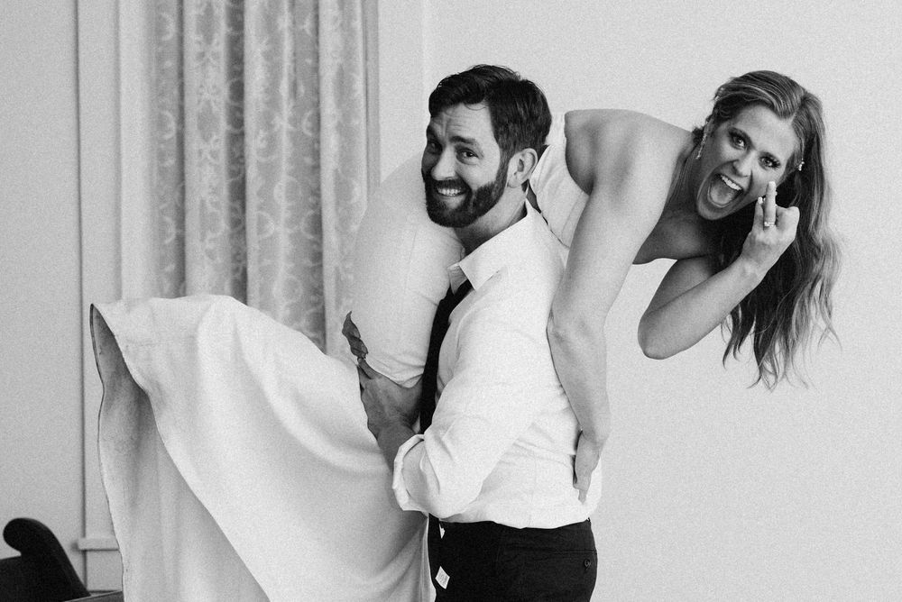 Black and white photo of groom carrying bride on his back, both smiling joyfully on their wedding day.