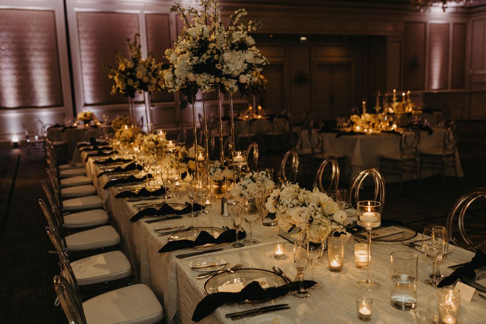 Elegant wedding reception with long tables, gold candelabras, white floral centerpieces, candlelight, and black napkins in a ballroom.