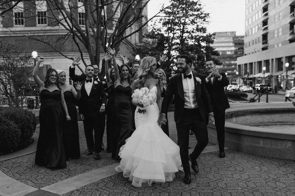 Black and white photo of a bride and groom walking with their wedding party outdoors near a fountain.