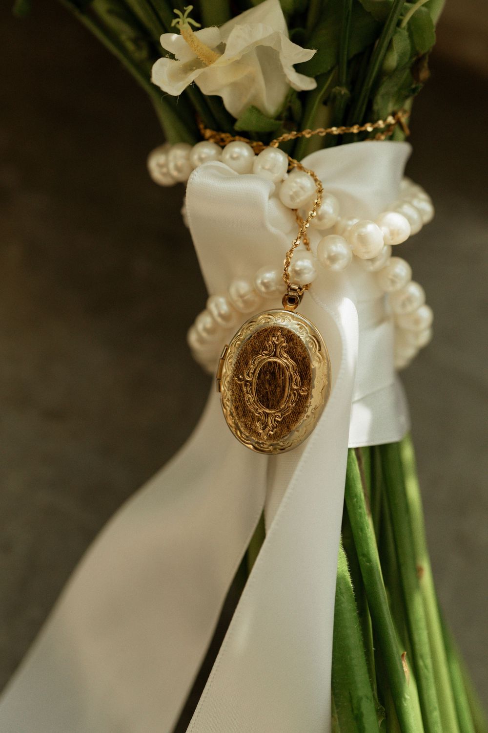 Gold locket pendant on pearl bracelet adorning a bridal bouquet with white ribbon and fresh flowers.
