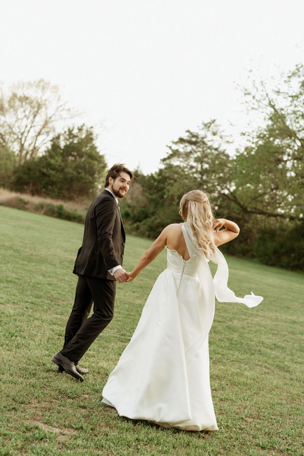 A smiling groom in a dark suit holds hands with a bride in a white gown as they walk across a green lawn.
