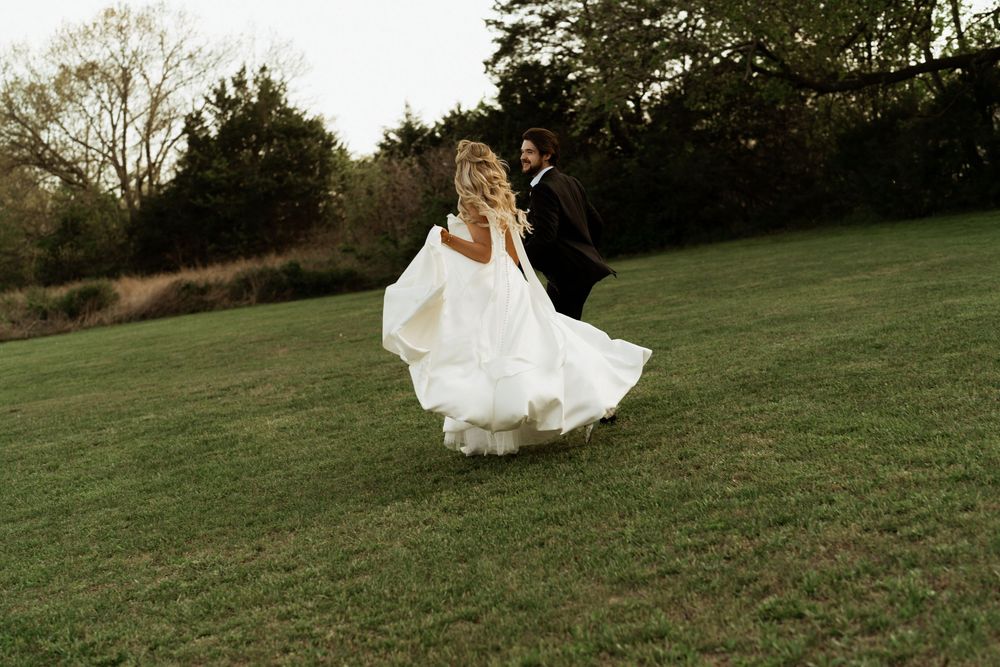 Bride in flowing white gown and groom in dark suit running across lush green lawn at outdoor wedding.