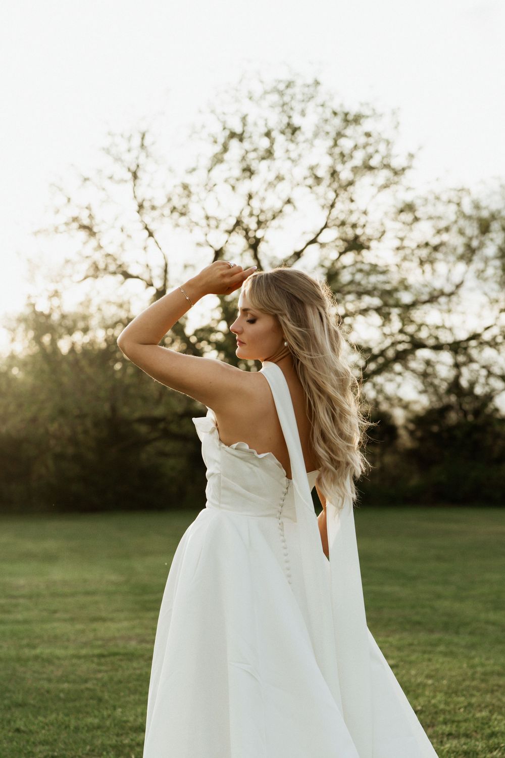 A bride in a white gown poses outdoors at golden hour, her long blonde hair flowing as she gazes away.