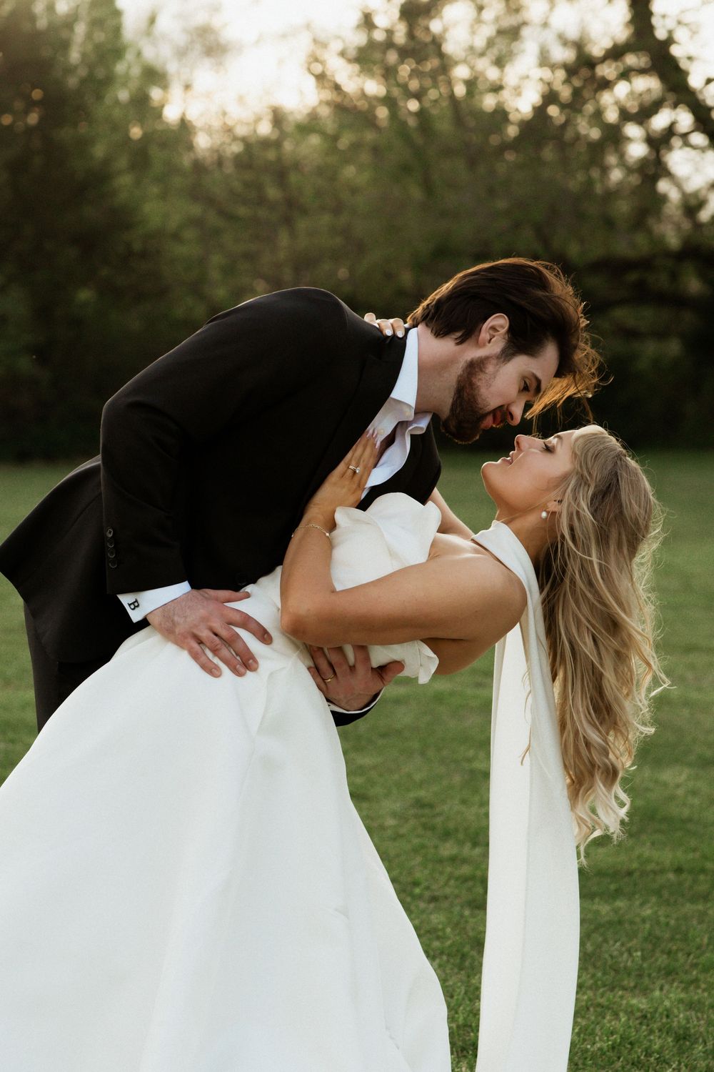 Groom in black tux dips smiling bride in white gown outdoors on green lawn with trees at golden hour.