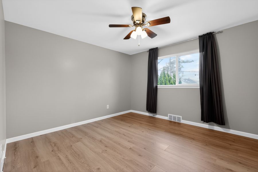 Empty bedroom with light wood floors, ceiling fan, gray walls, and dark curtains framing a window.