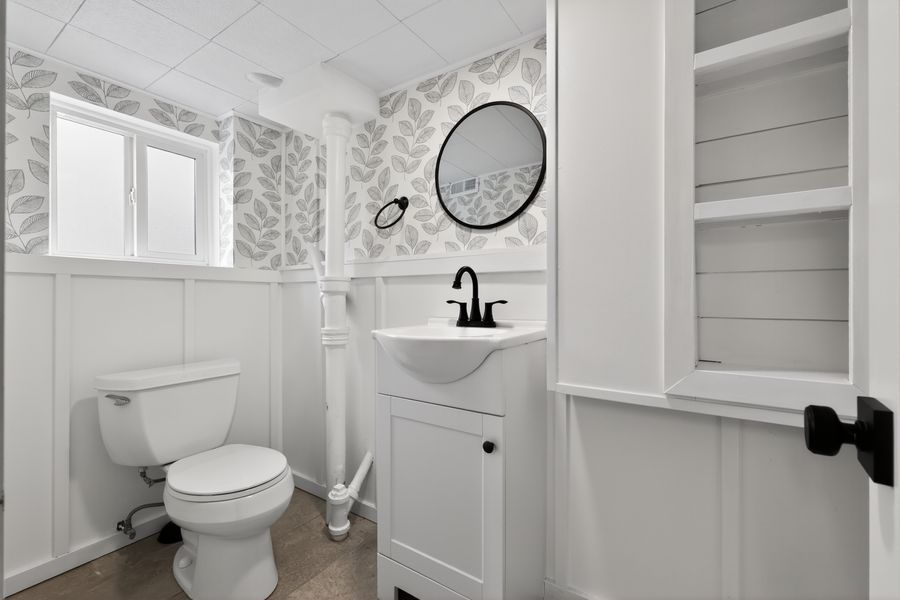 White bathroom with round mirror, pedestal sink, black fixtures, wainscoting, and patterned wallpaper above.