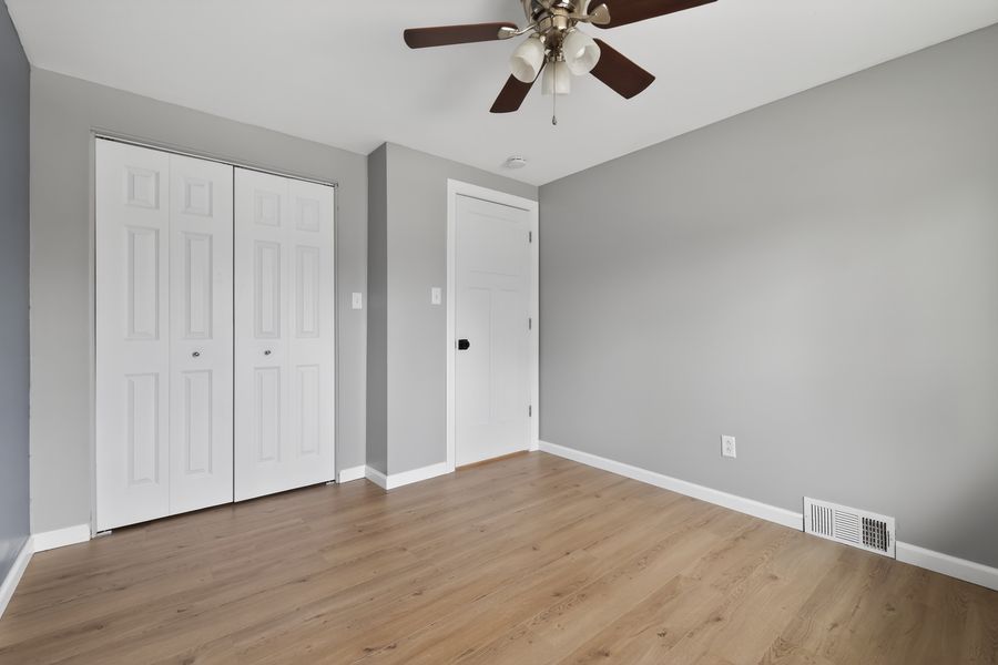Empty bedroom with ceiling fan, hardwood floors, white closet doors, and neutral gray walls.