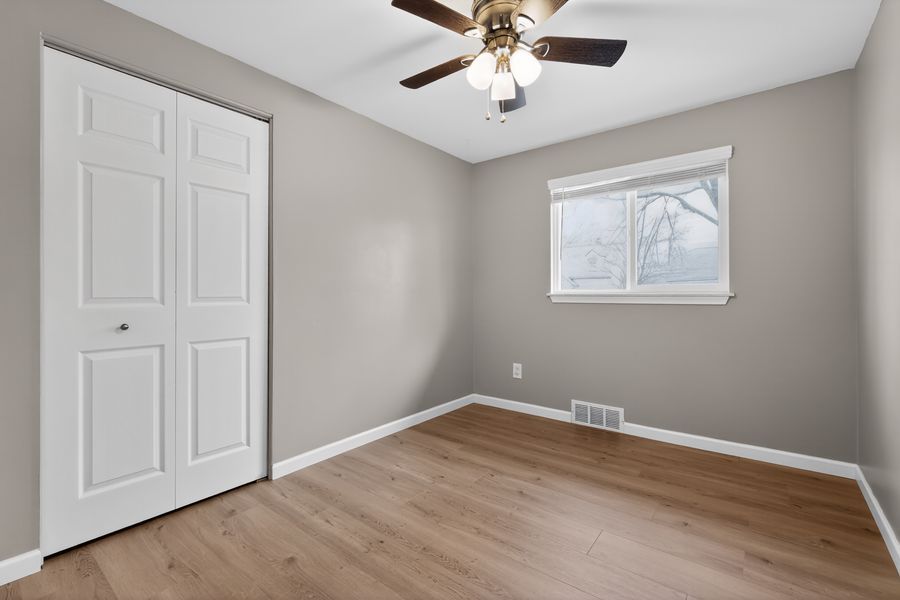 Empty bedroom with light wood floors, gray walls, white bifold closet door, ceiling fan, and a window with natural light.