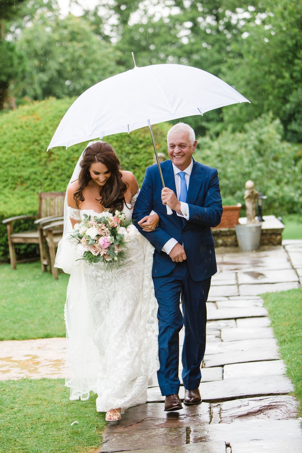 Bride in lace gown holding pink bouquet walks with suited man under white umbrella on rainy garden wedding day.