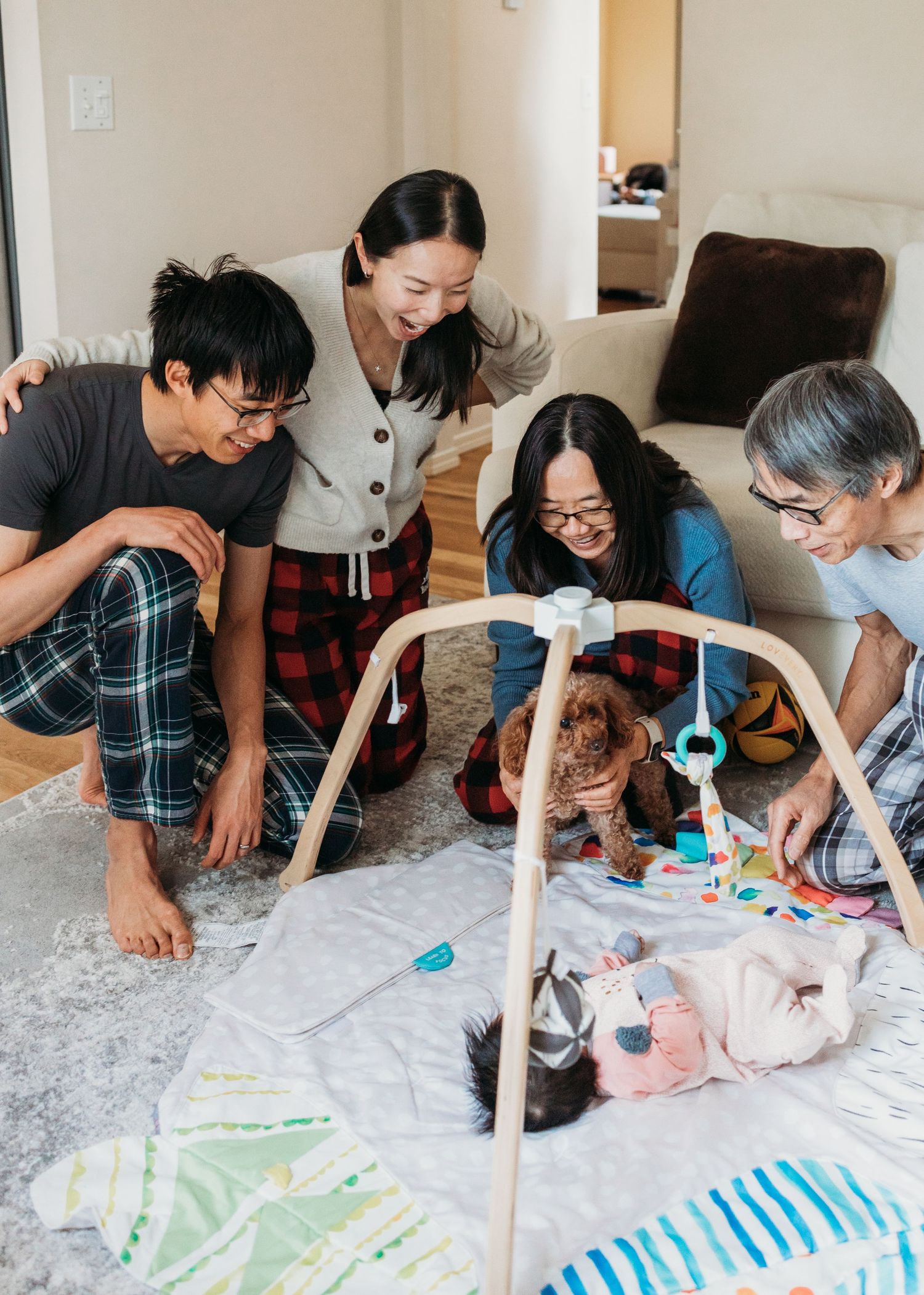 Asian family gathered around a baby play gym on the floor, smiling and bonding with a small dog nearby.