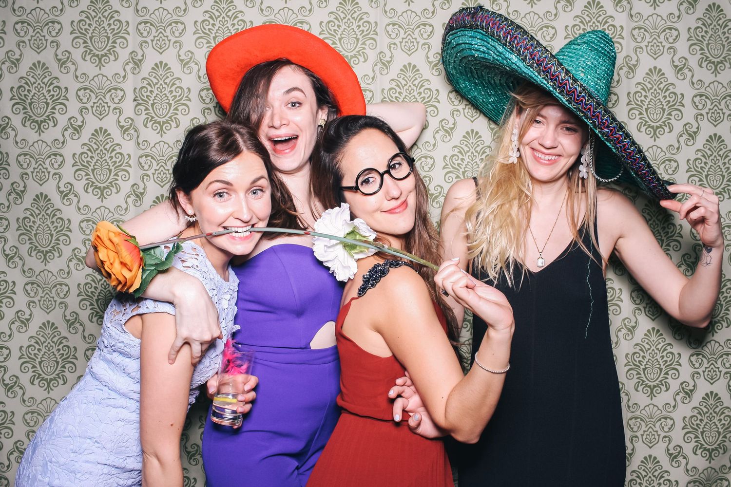 Four young women posing playfully in a photo booth wearing colorful hats and fun props against a floral wallpaper backdrop.