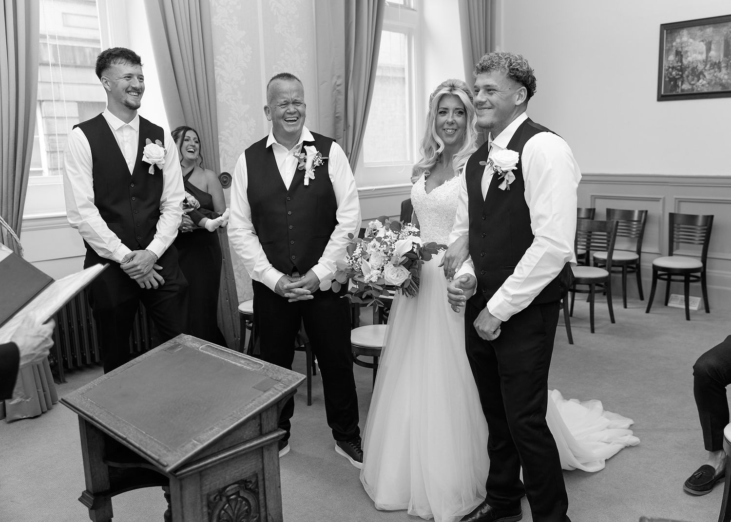 Black and white wedding ceremony photo showing bride with bouquet and groom smiling with guests in elegant room.