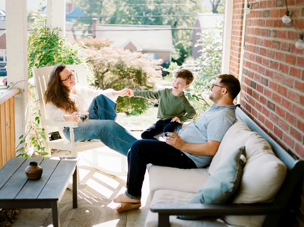 Family of three laughing and relaxing together on a cozy covered front porch with brick walls.