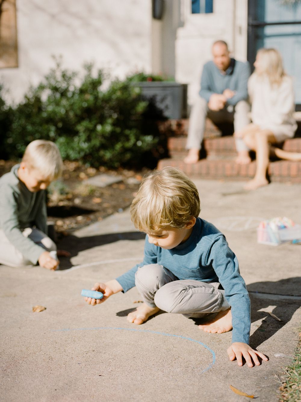 Two young children draw with sidewalk chalk on a patio while parents relax on steps in the background.