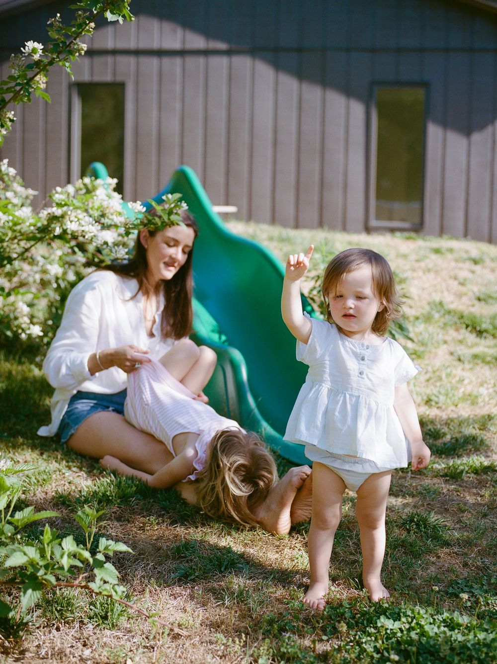Woman in floral crown sits on grass with two young children near a green slide and blooming bush on sunny day.