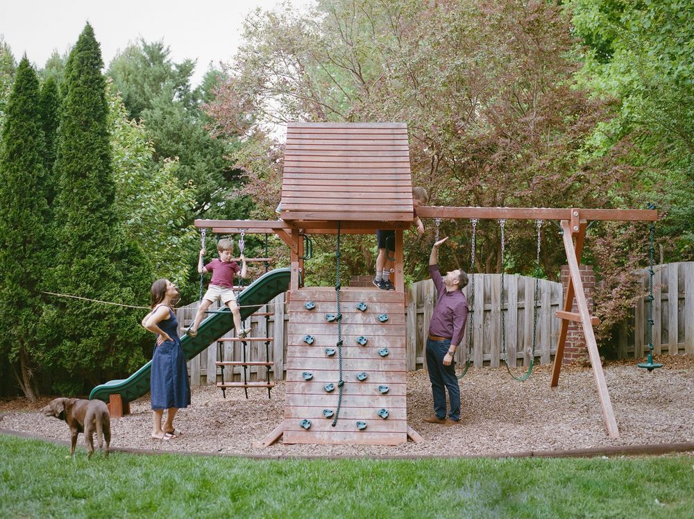 Family playing on a wooden backyard playset with climbing wall, swings, and slide, surrounded by lush green trees.