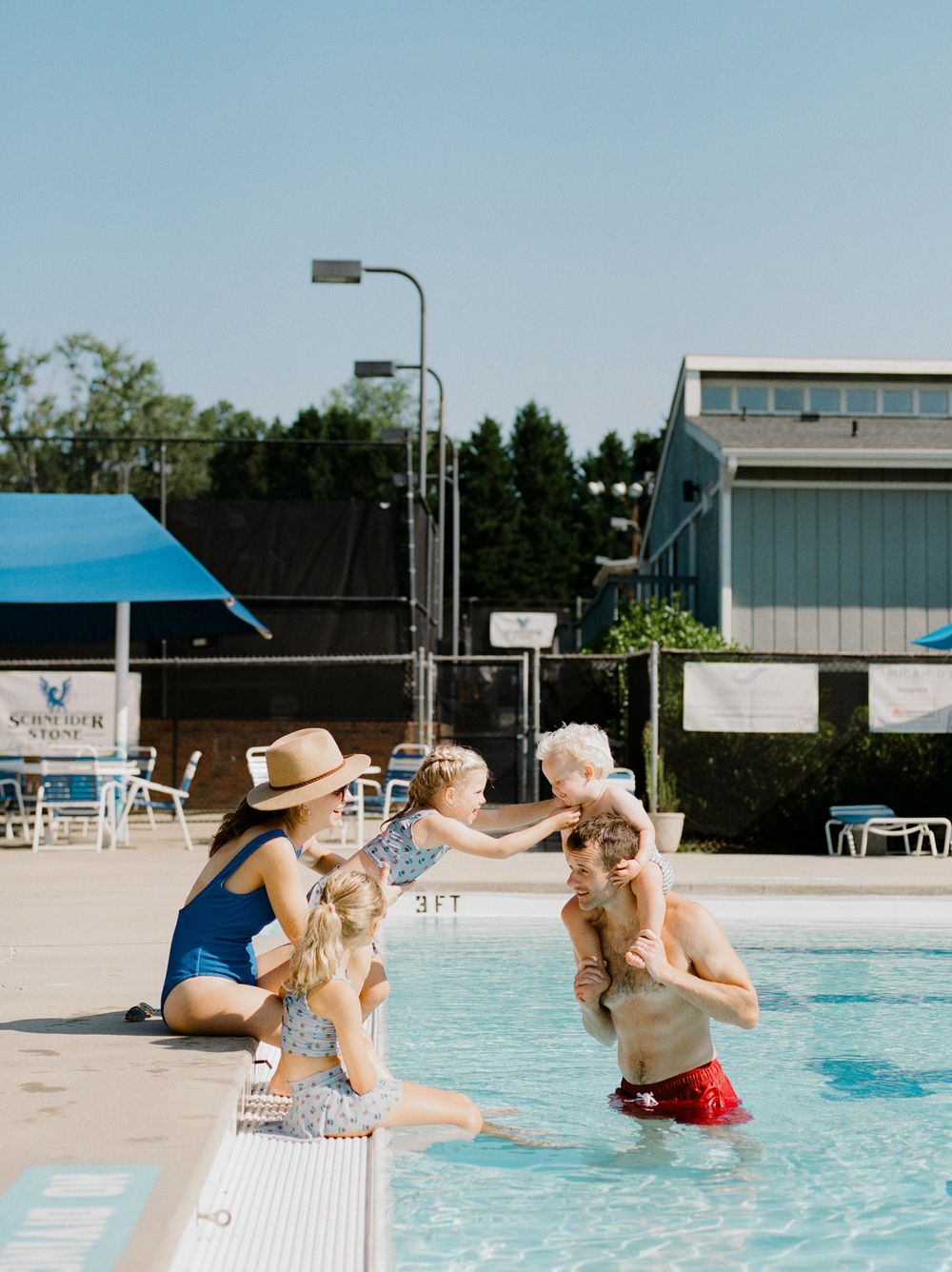 Family enjoying a sunny day at an outdoor pool, with children splashing and playing in the shallow end near the pool edge.
