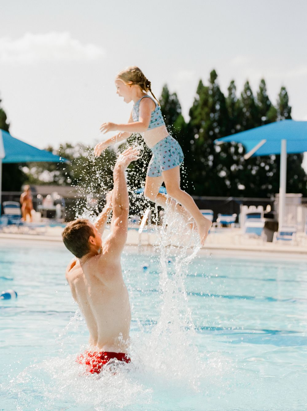 Man tossing young girl into the air at an outdoor swimming pool on a sunny day with blue umbrellas in background.