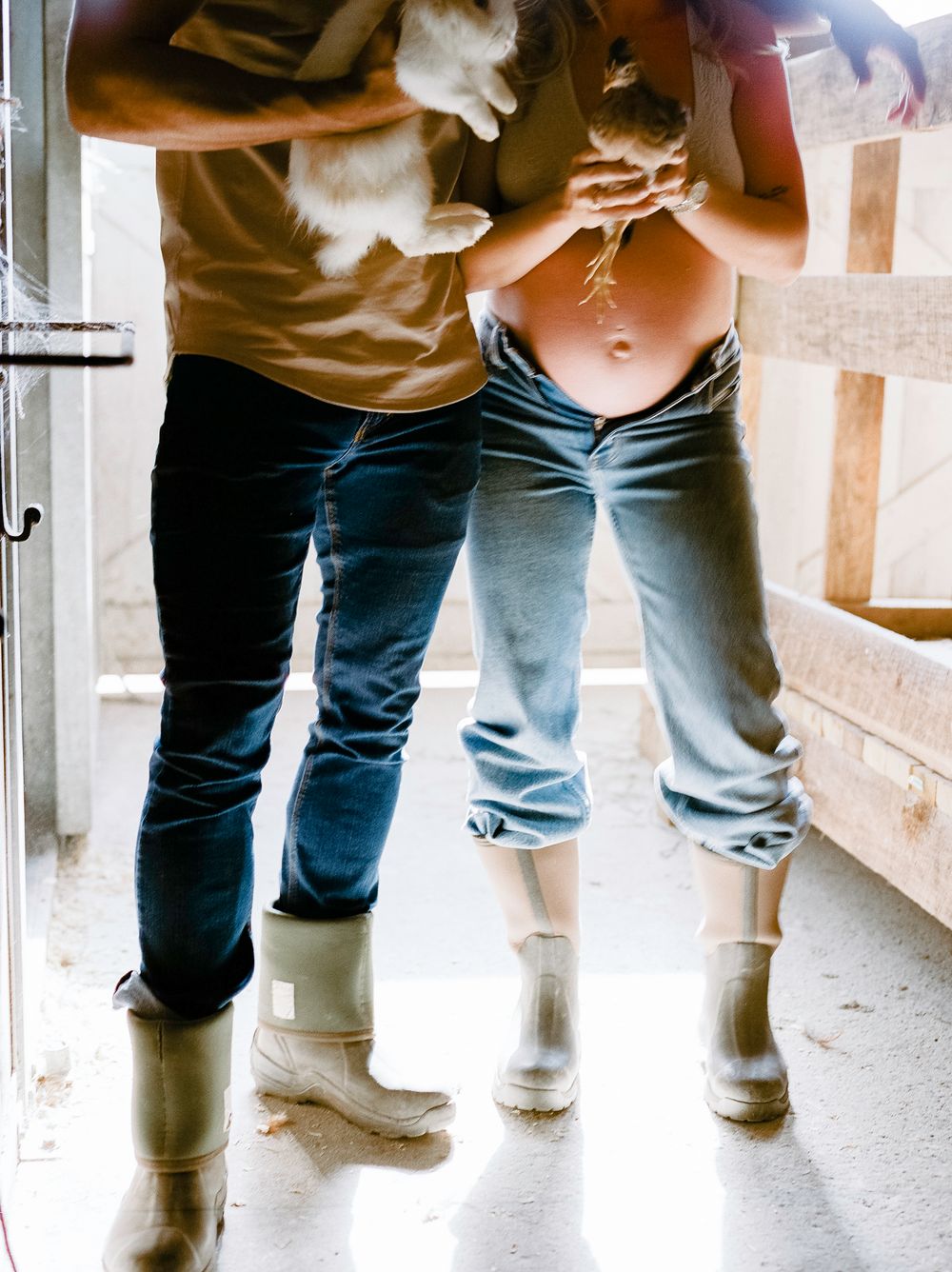 Couple in jeans and rain boots holding baby animals, with pregnant belly visible, standing in a barn doorway.