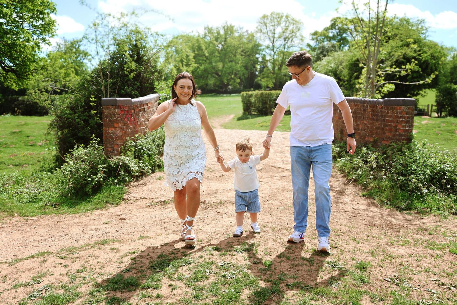 Family of three walking hand-in-hand outdoors, parents swinging toddler between them on a sunny day in a lush green park.