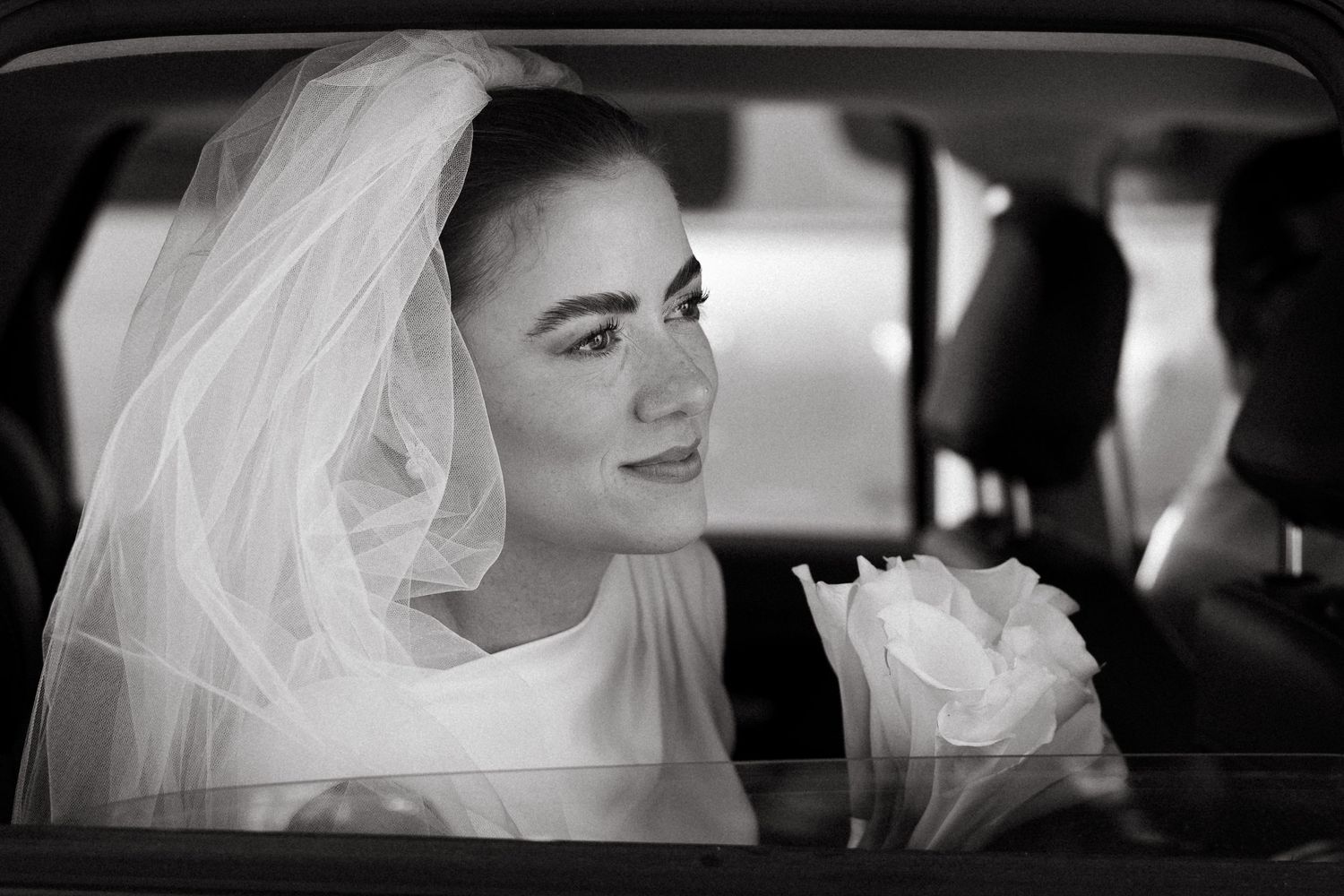 Black and white portrait of a beautiful bride wearing a veil sitting in a car on her wedding day.