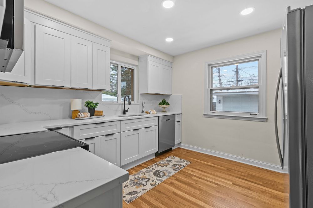 Modern white kitchen with marble countertops, stainless steel appliances, hardwood floors, and bright natural light.