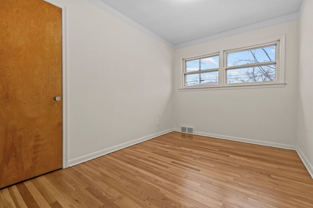 Bright empty bedroom with light hardwood floors, white walls, and a large window providing natural light.
