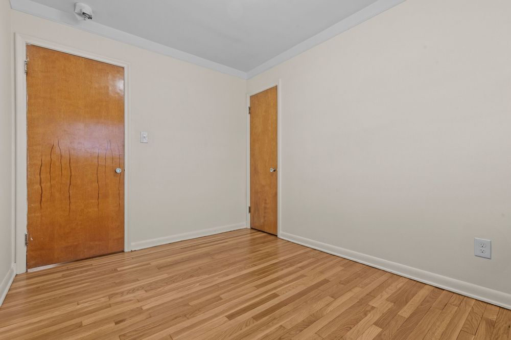 Empty room with light hardwood floors, cream walls, crown molding, and two wooden doors in a residential apartment.