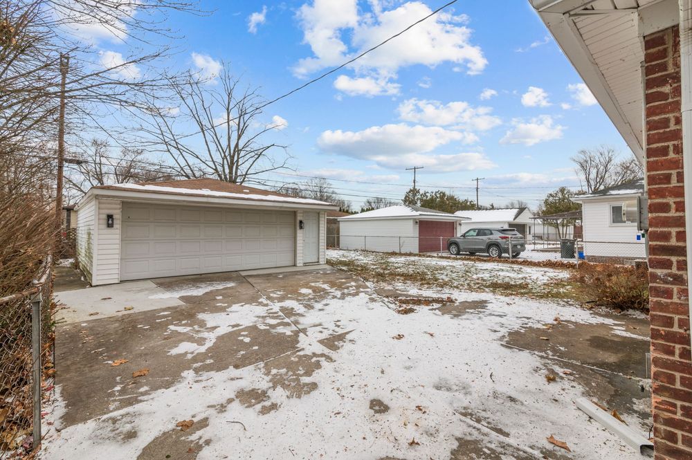 Snow-dusted backyard with detached white garage, concrete driveway, and neighboring homes under a partly cloudy winter sky.