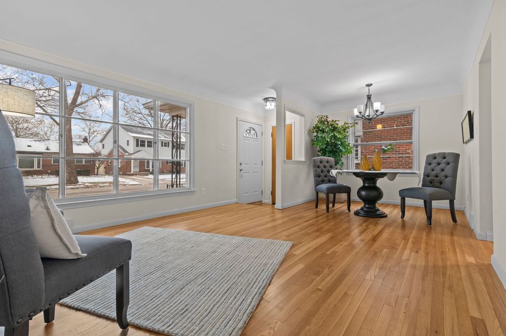 Bright living and dining area with hardwood floors, large windows, chandelier, and modern gray chairs on a striped rug.