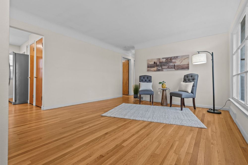 Bright living room with hardwood floors, two blue chairs, a gray area rug, and a floor lamp against white walls.
